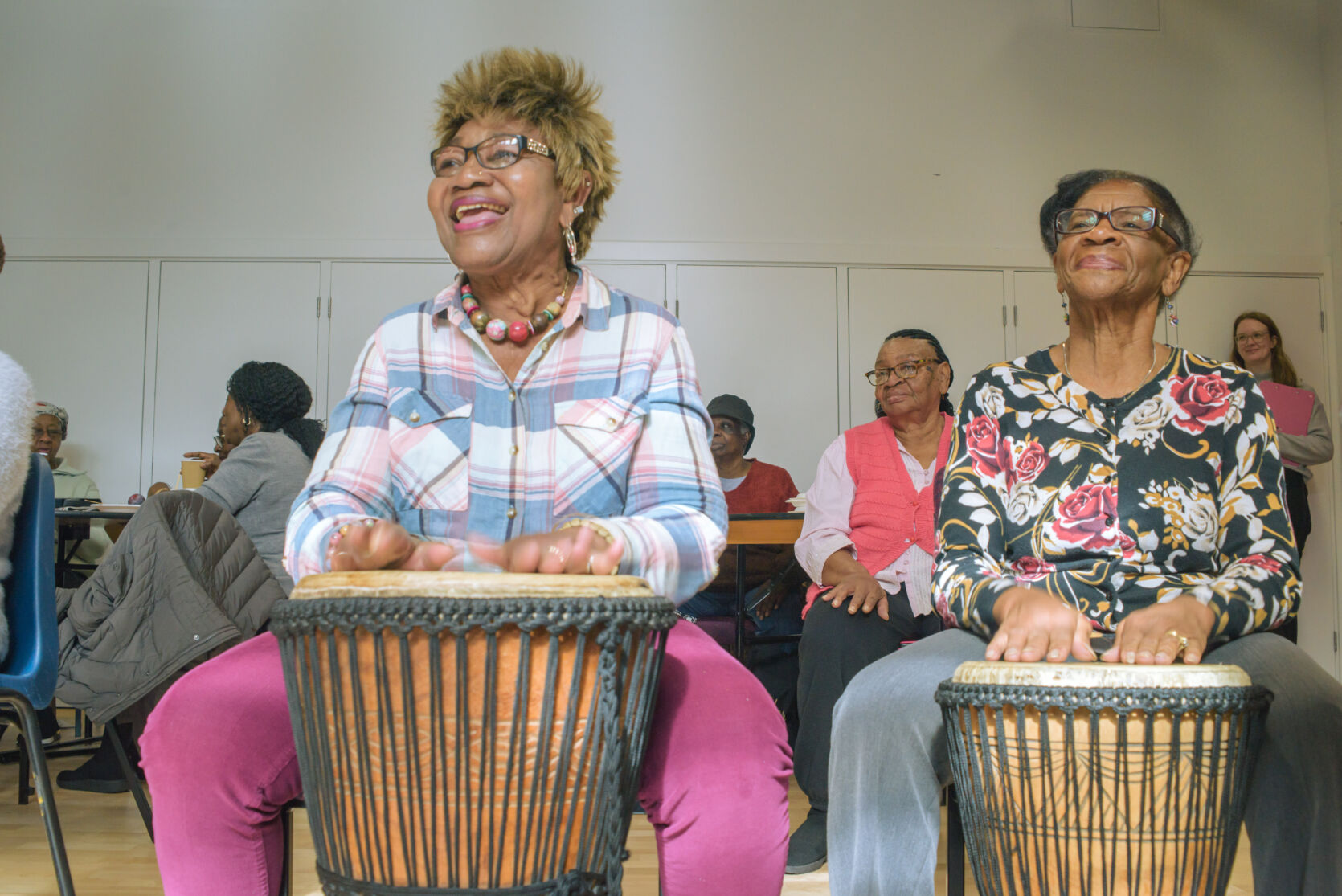Two people sitting with drums in front of them, ready to play. In the background are other people also sitting but without drums.