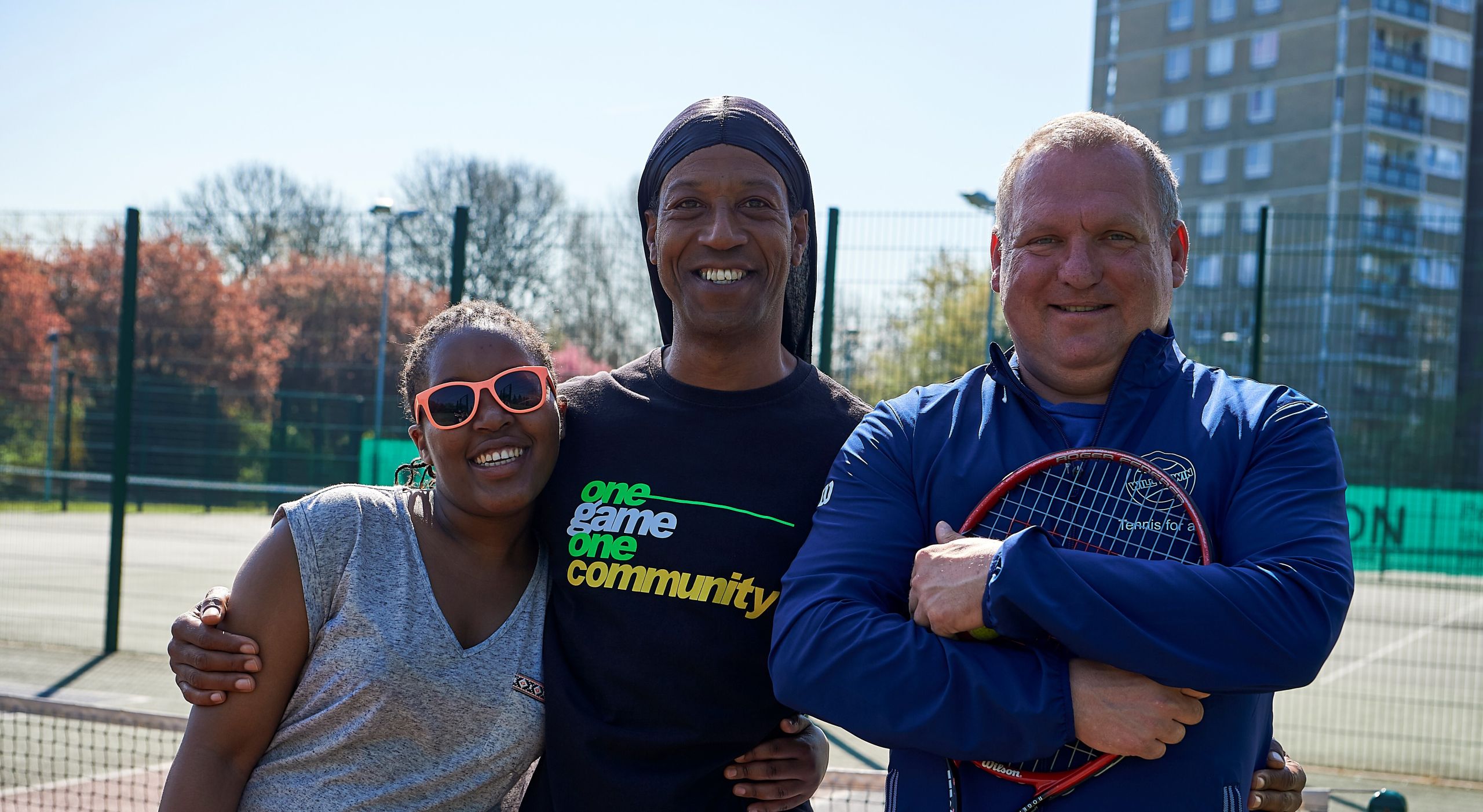 Three people standing together on a tennis court - two are older and one is younger. The oldest person is holding a tennis racquet and the youngest has brightly coloured sunglasses. In the background can be seen a group of trees and a tower block.