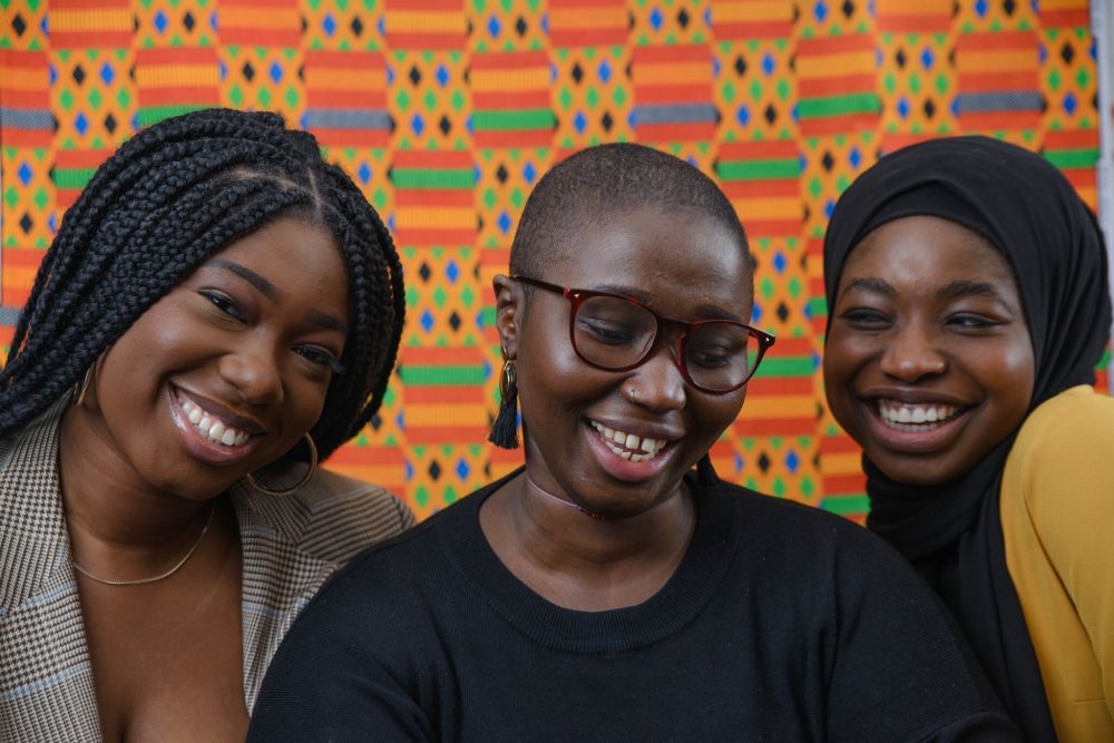 Three Black woman smile to camera in front of a patterned multicoloured background
