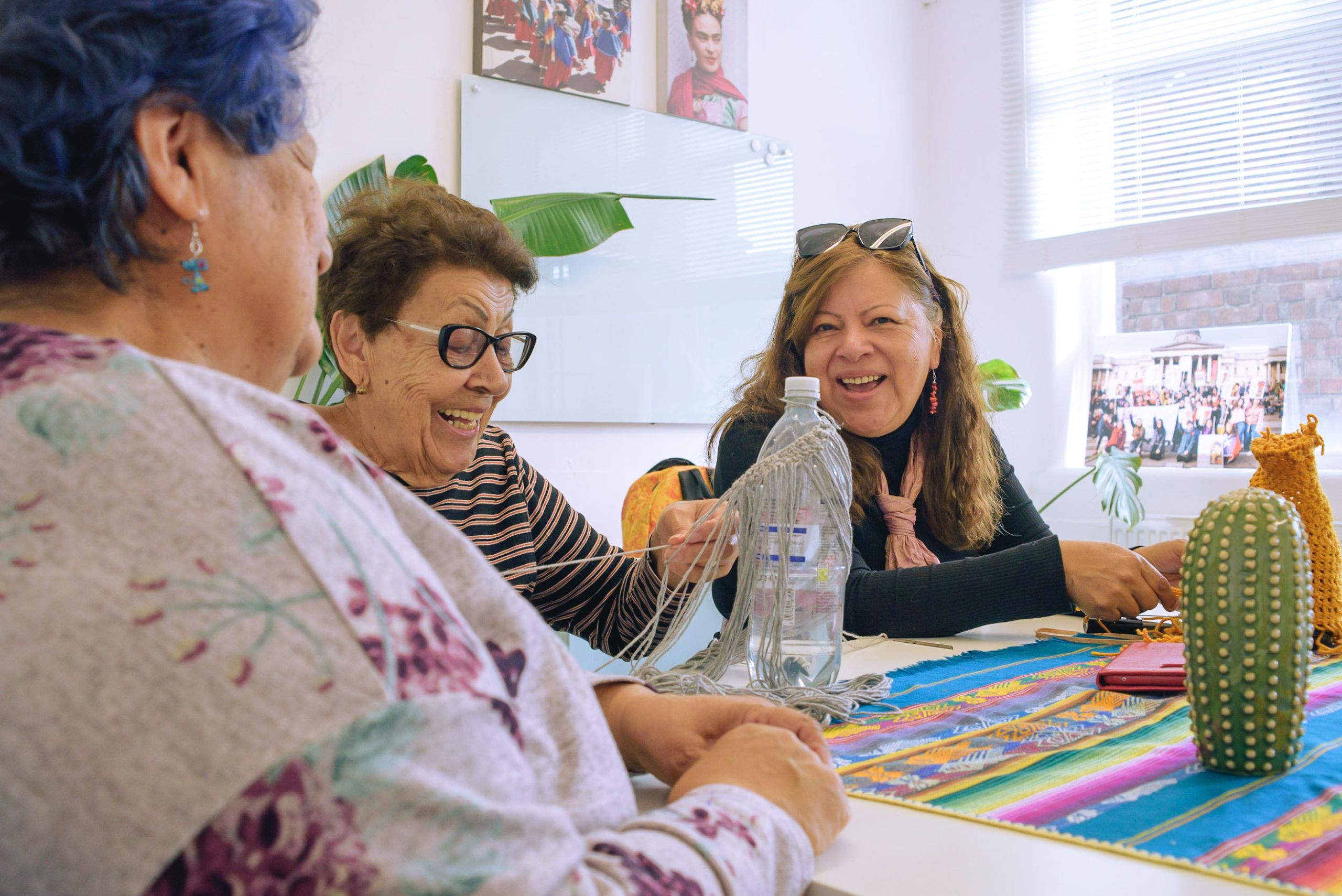 Three ladies sitting together at a table, taking part in craft activities. They are happily chatting to one another as they work. There's a cactus in front of them and some plants behind them.