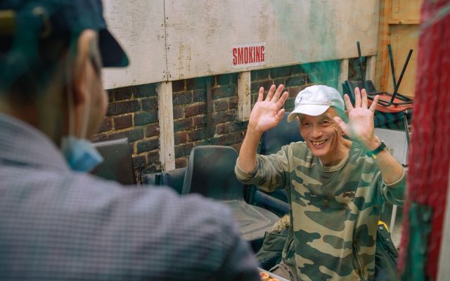 An older man sitting at a table with his hands raised and a smile on his face. He's looking at another person standing just in front of the camera but we can only see the back of him.