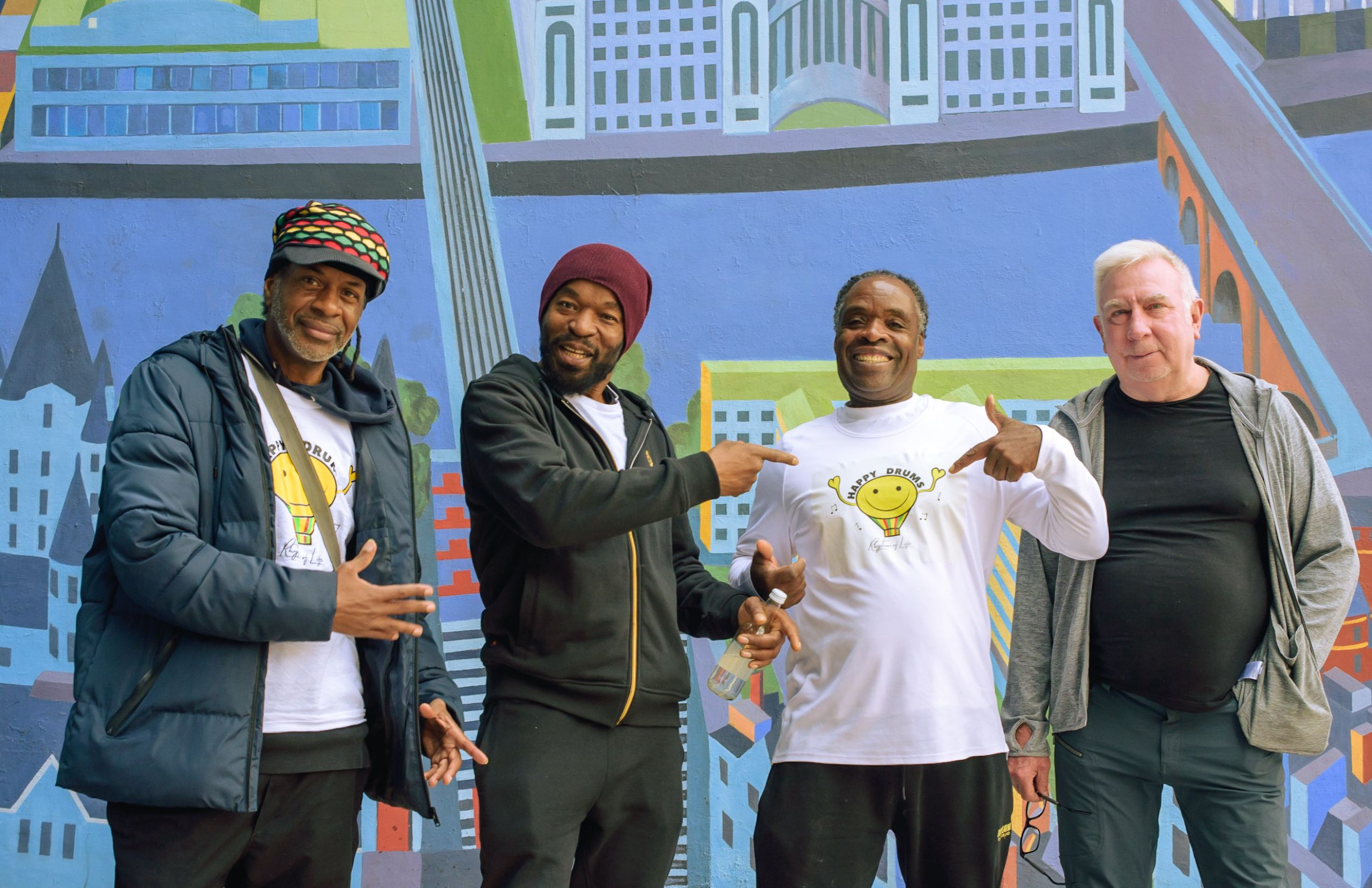 Four people standing together in a line in front of colourful wall mural. Three of them are pointing to one of the people's t-shirts that says 'Happy Drums' on it.