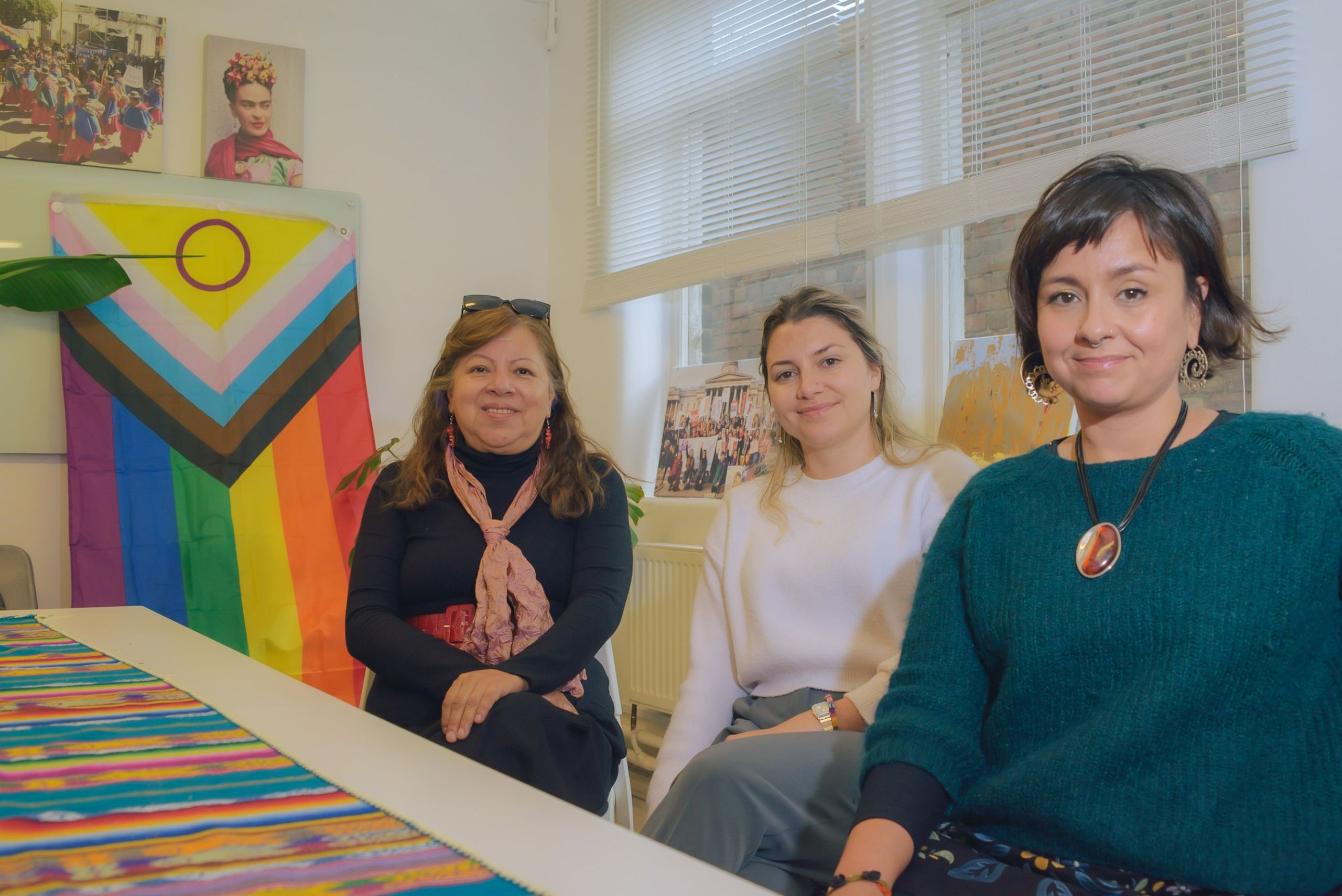 Three women sitting at a table, looking straight at the camera. In the background is a brightly coloured flag and some pictures on the wall.