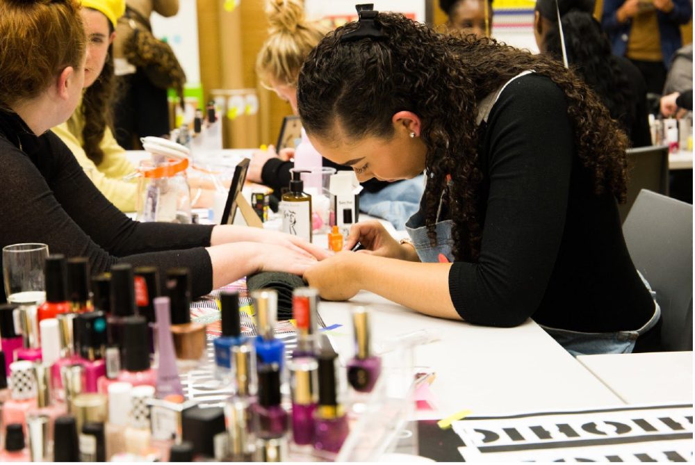 A young person receiving treatment on their nails in a nail bar. In the foreground are lots of different bottles of nail varnish and other young people can be seen in the distant background too.