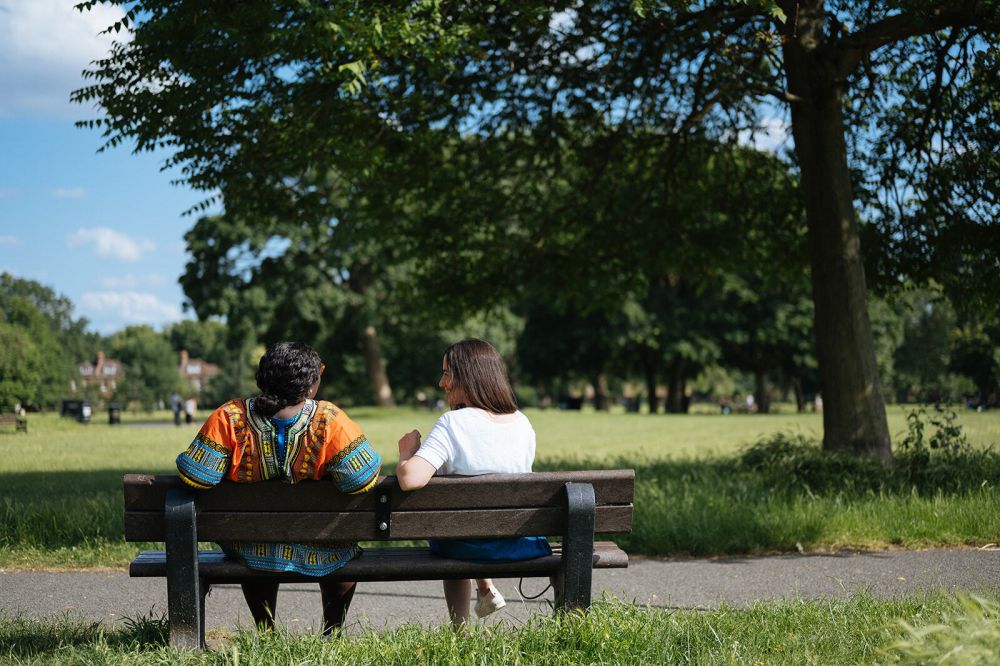 Two people sitting on a park bench, seen from behind. In the background is a large area of grass and a range of different trees. The nearest tree almost fills the whole background.