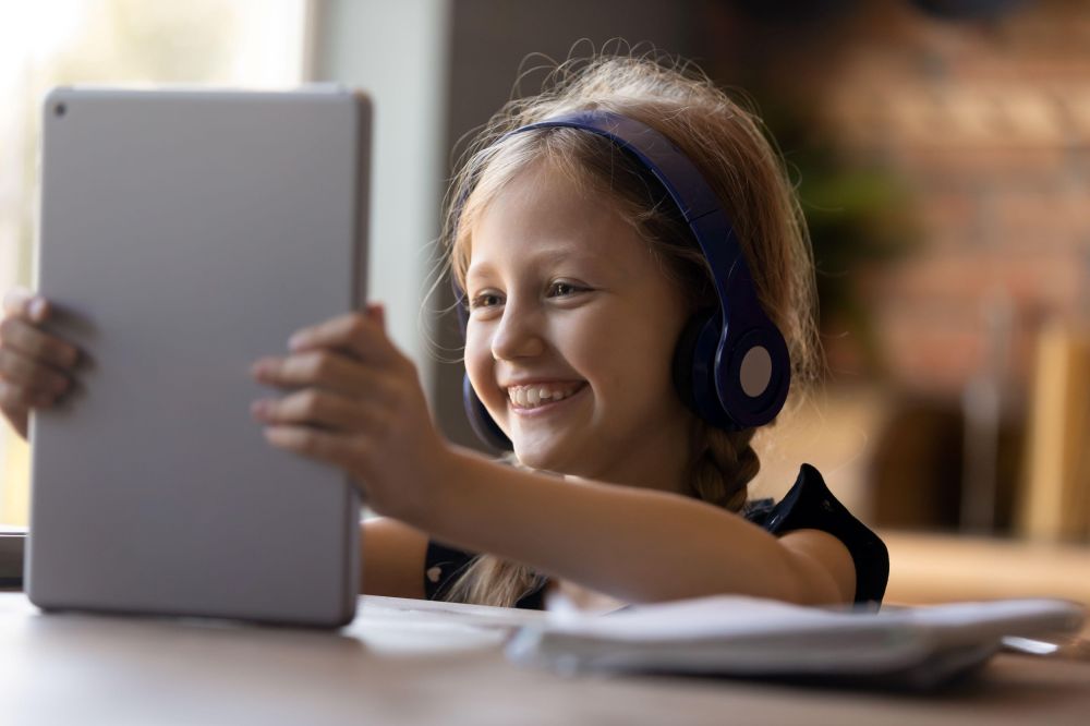 A young girl is sitting at a table, using a tablet device. She is wearing large, over-ear headphones and there are some sheets of paper in front of her. She has a big smile on here face.