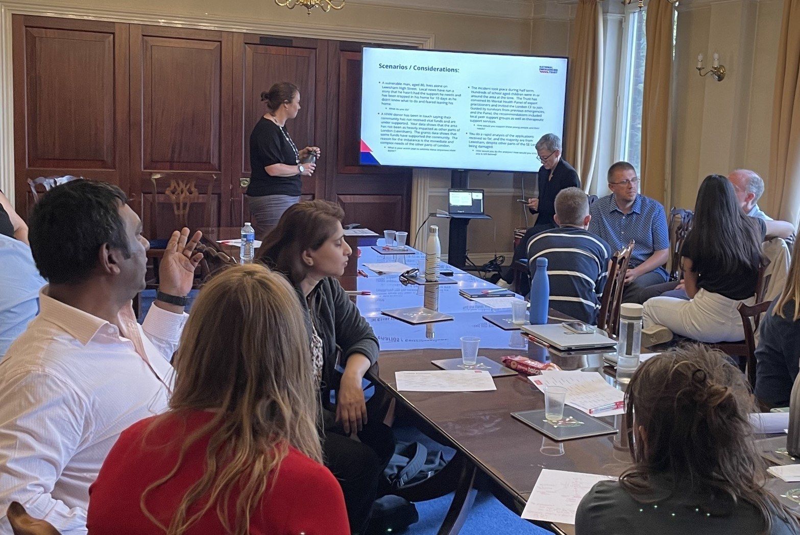 Several groups of people are gathered around a long boardroom table, listening to a presentation by Helen Killingley from The National Emergencies Trust.