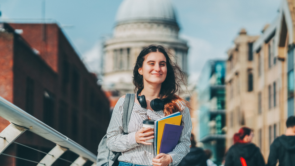 A young person walking across a bridge in London. They are carrying study books and a cup of coffee, with large over-ear headphones hanging around their neck. In the background you can see buildings, including St Paul's Cathedral.