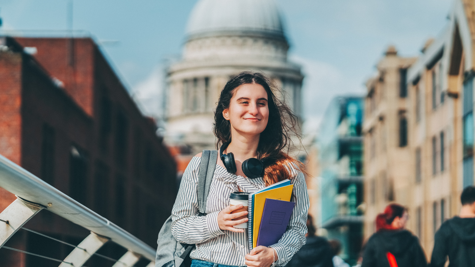 A young person walking across a bridge in London. They are carrying study books and a cup of coffee, with large over-ear headphones hanging around their neck. In the background you can see buildings, including St Paul's Cathedral.