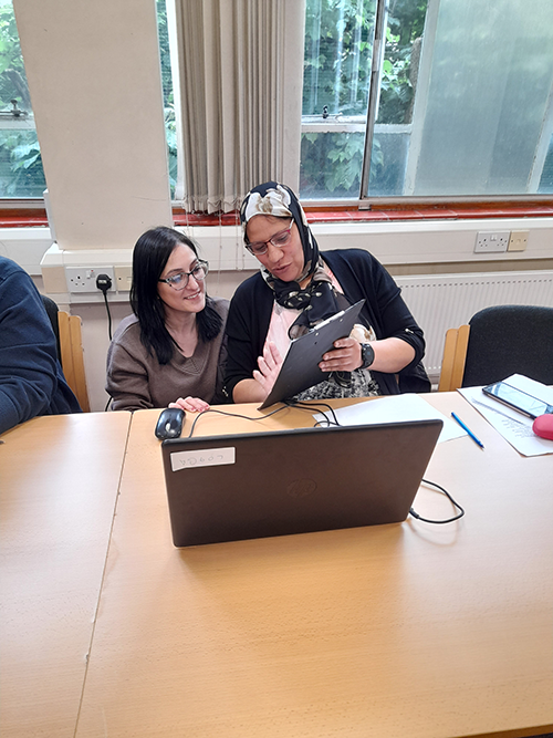 Two women sit behind a laptop at a table. One woman holds a clipboard and points something out to the other woman who is crouching on the floor.