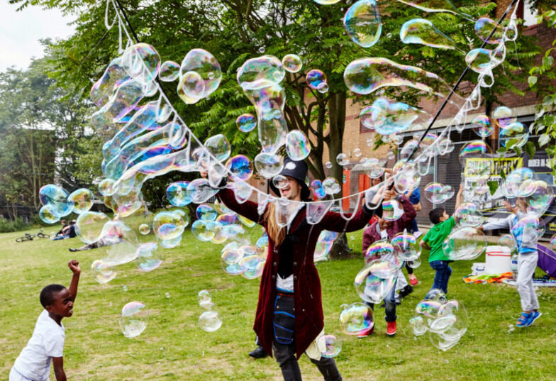 A man dressed in a velvet tailcoat, tartan trousers and a top hat holds up a large string on two sticks, through which he blows bubbles. Children around him play in the bubbles.