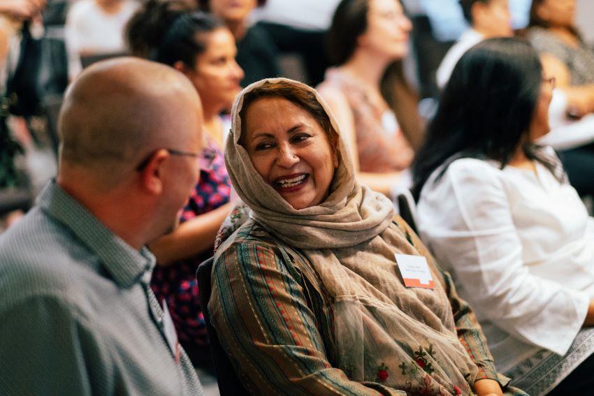 Two people sitting together, talking, in a crowded audience. Both are wearing name badges and they are enjoying their conversation together.