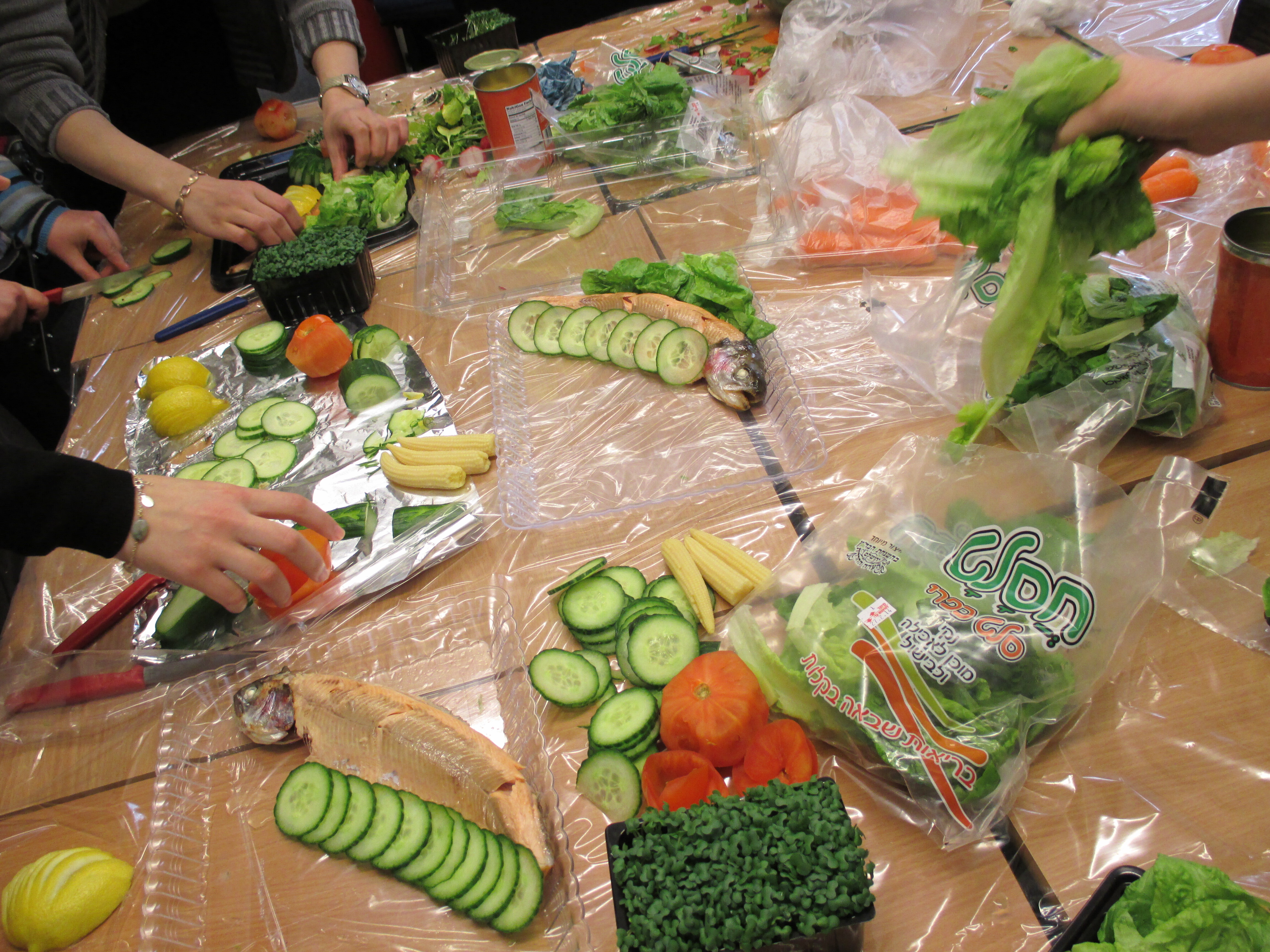 A table is covered with plastic sandwich bags and chopped salad ingredients. Some young girls' hands are in shot chopping the vegetables and putting them into bags.
