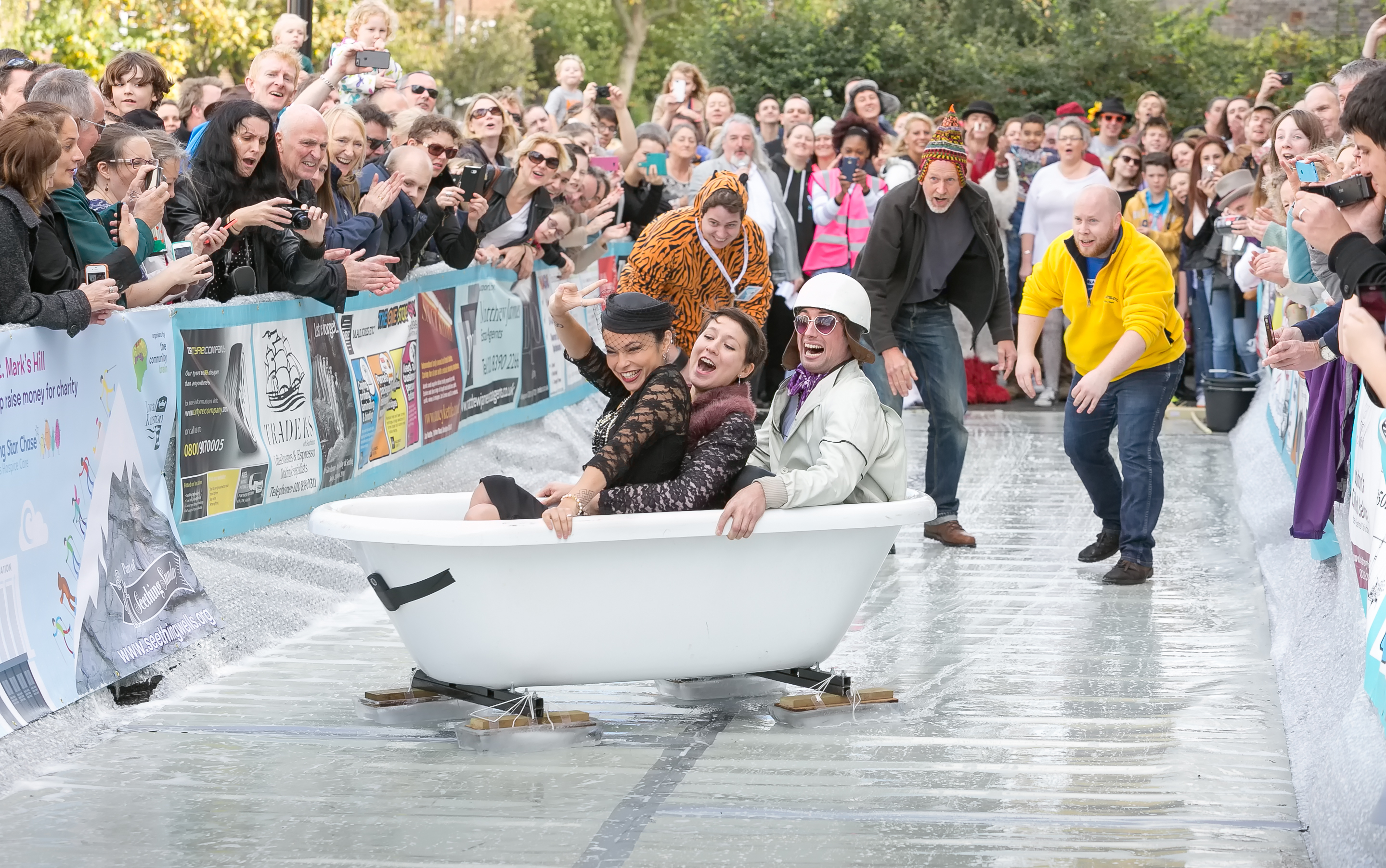 Two women and a man are in a bathtub in fancy dress smiling and laughing. They slide along a slippery surface as an onlooking crowd takes photos. Behind them, three men who have pushed the bathtub look on.