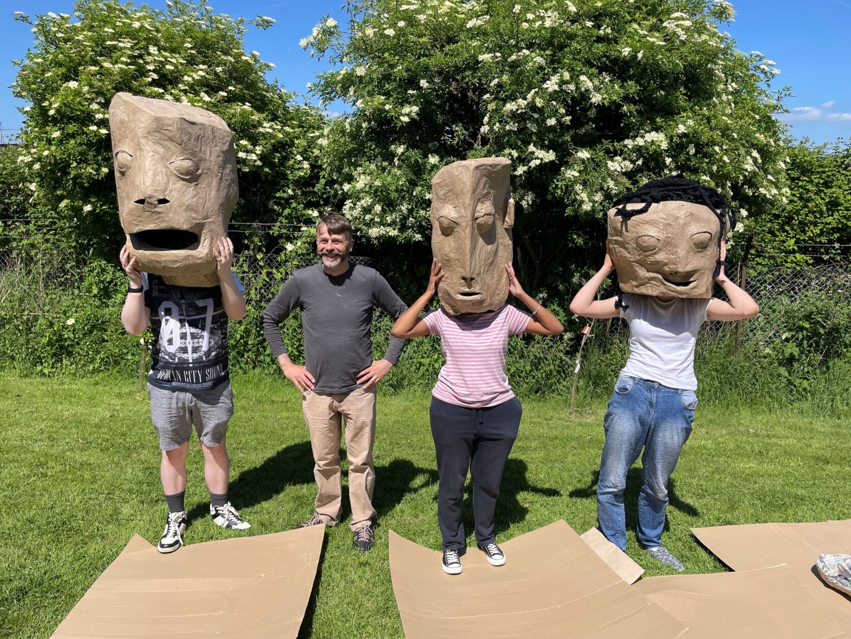 A group of four people standing in a line in a park. Three of them have large masks on their heads, featuring strange and unusual faces.