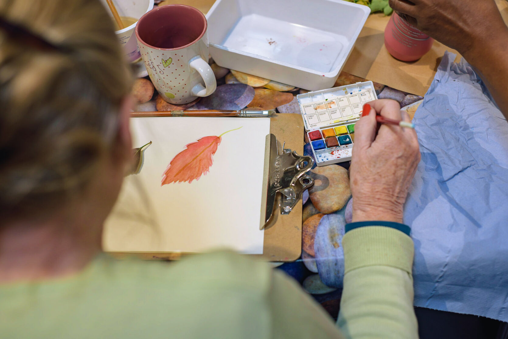 A person seen from behind and overhead, painting a picture of a leaf. They are surrounded by art supplies and a mug.