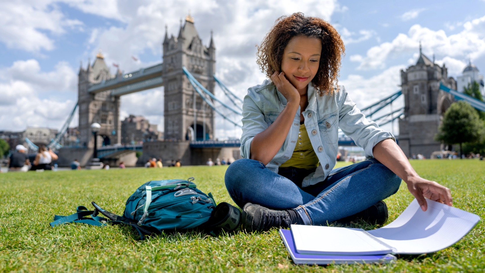 A young person sitting on grass with study books in front of them and a bag next to them. Behind them you can see Tower Bridge in London as well as blue sky and white fluffy clouds.