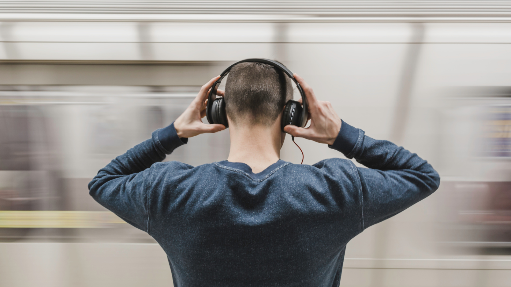 A young person, seen from behind and wearing large over-ear headphones. Behind them can be seen a moving train.