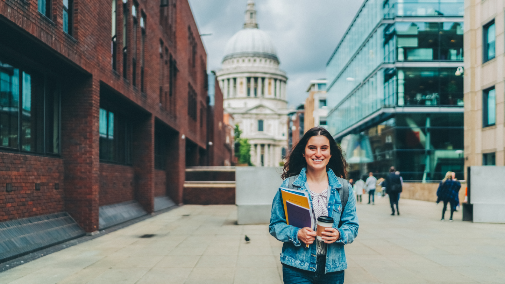 A young person holding study books and a cup of coffee. Either side of them are two rows of office buildings and in the background is St Paul's Cathedral in London.