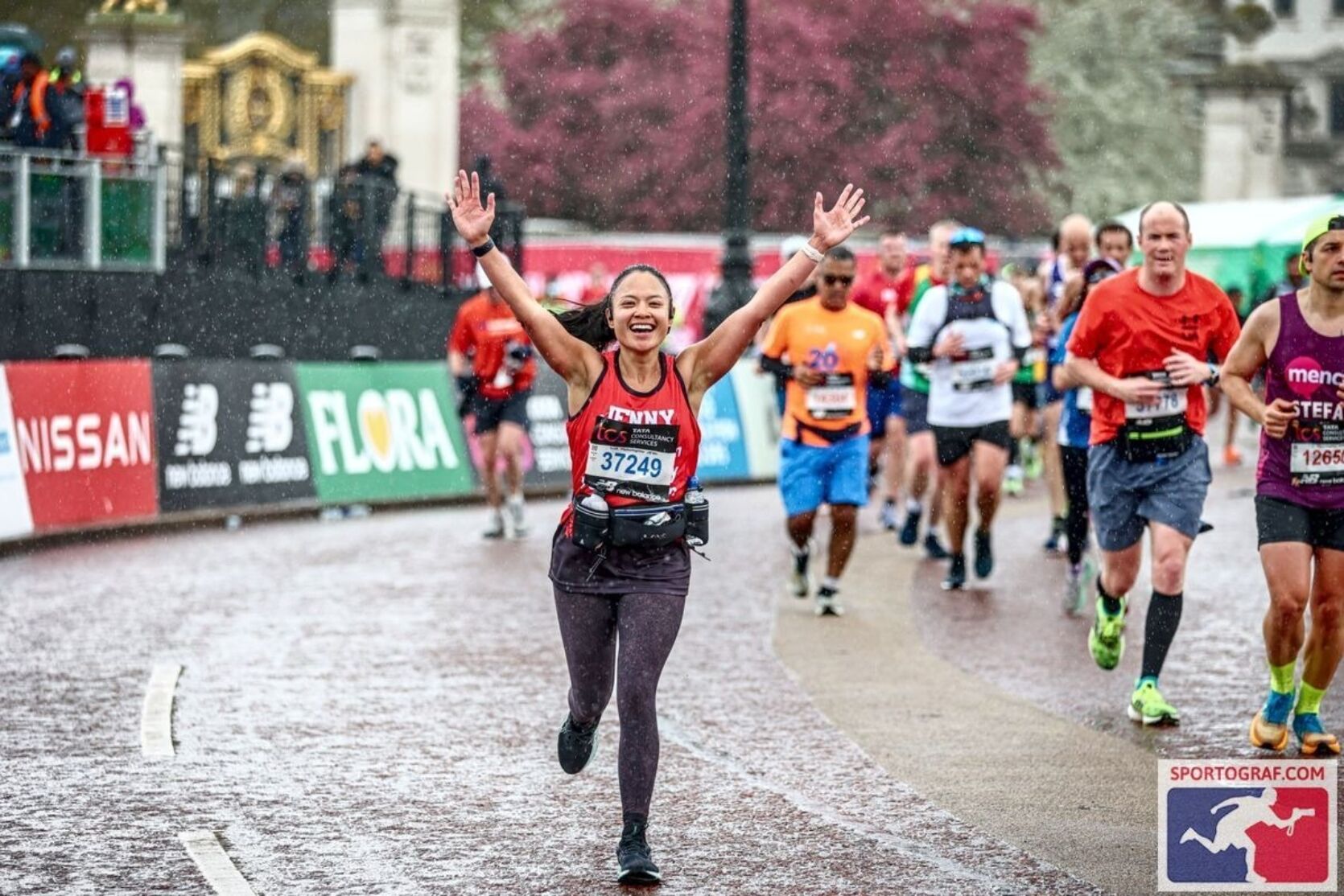 A marathon runner, wearing a red The London Community Foundation running vest, raises their arms as they run towards the camera. The name on their vest is 'Jenny'. Behind them and to the right of the runner are many more marathon runners taking part, wearing a range of different coloured vests and shirts.