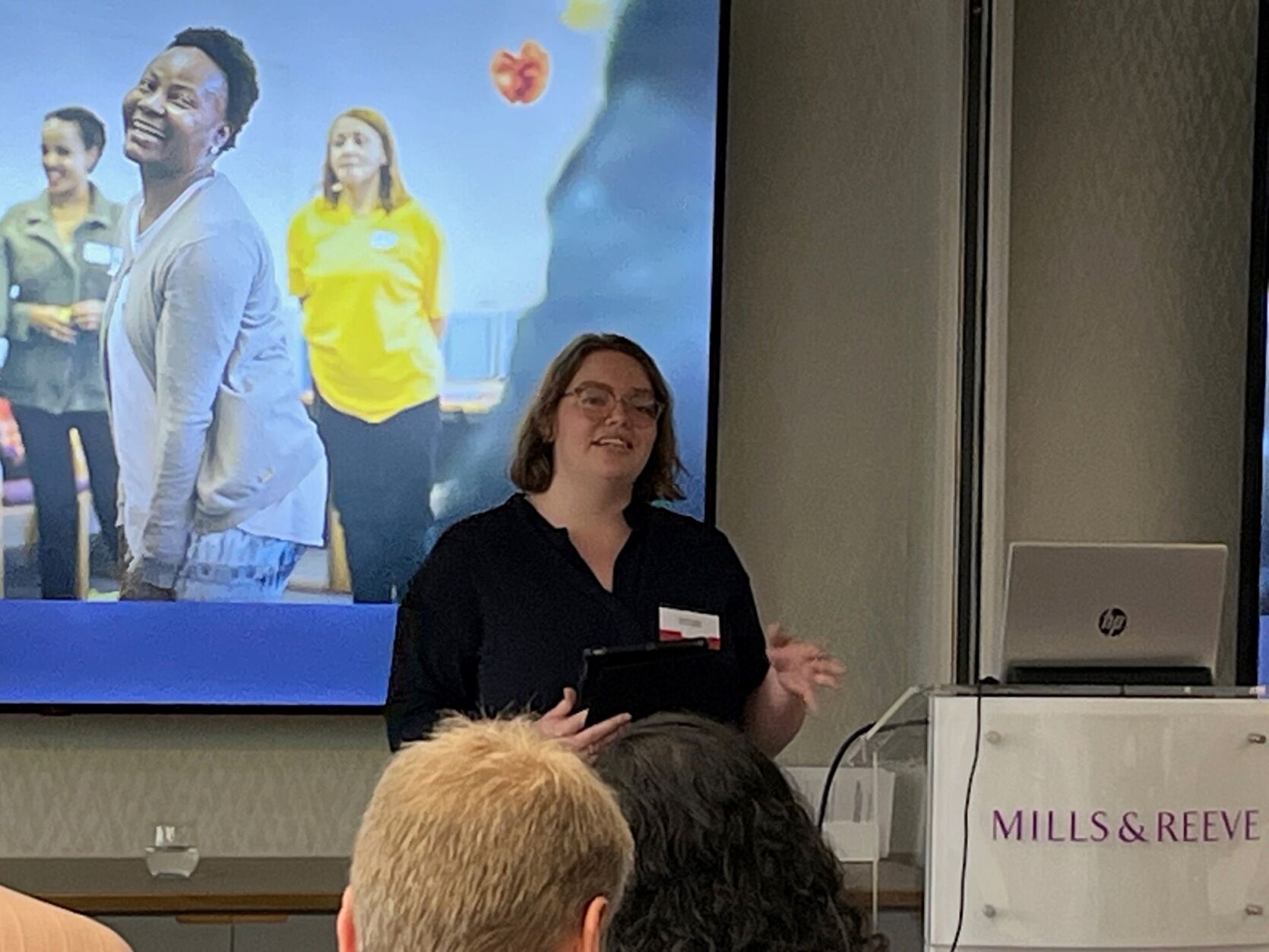 A person presenting at the Women's Fund for London event. In foreground is a podium with the words 'Mills and Reeve' on it and on the slides in the background is a group of young people.