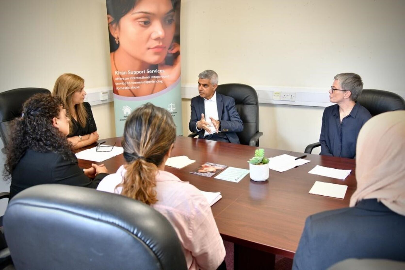 Members of the team from Kiran Support Services sitting around a boardroom table with the Mayor of London, Sadiq Khan, and The London Community Foundation CEO Kate Markey. In the background is a pull-up banner with a woman's face on it.