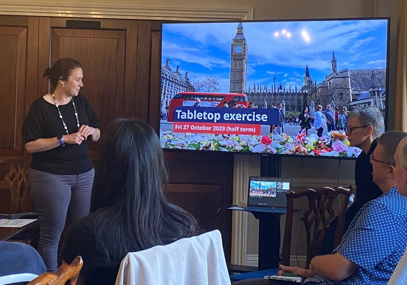 Helen Killingley from The National Emergencies Trust is presenting to a meeting. In the background can be seen four members of the audience (viewed from behind) and on the slides is a picture of Parliament Square with Big Ben and a red London bus visible too.