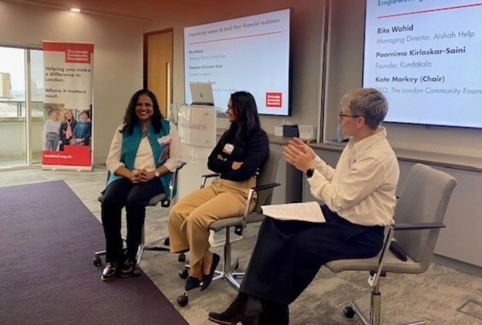 Panellists sharing their experiences at the Women's Fund for London event with CEO Kate Markey - behind them is a large screen, a podium with a laptop on it and a London Community Foundation pull-up banner.