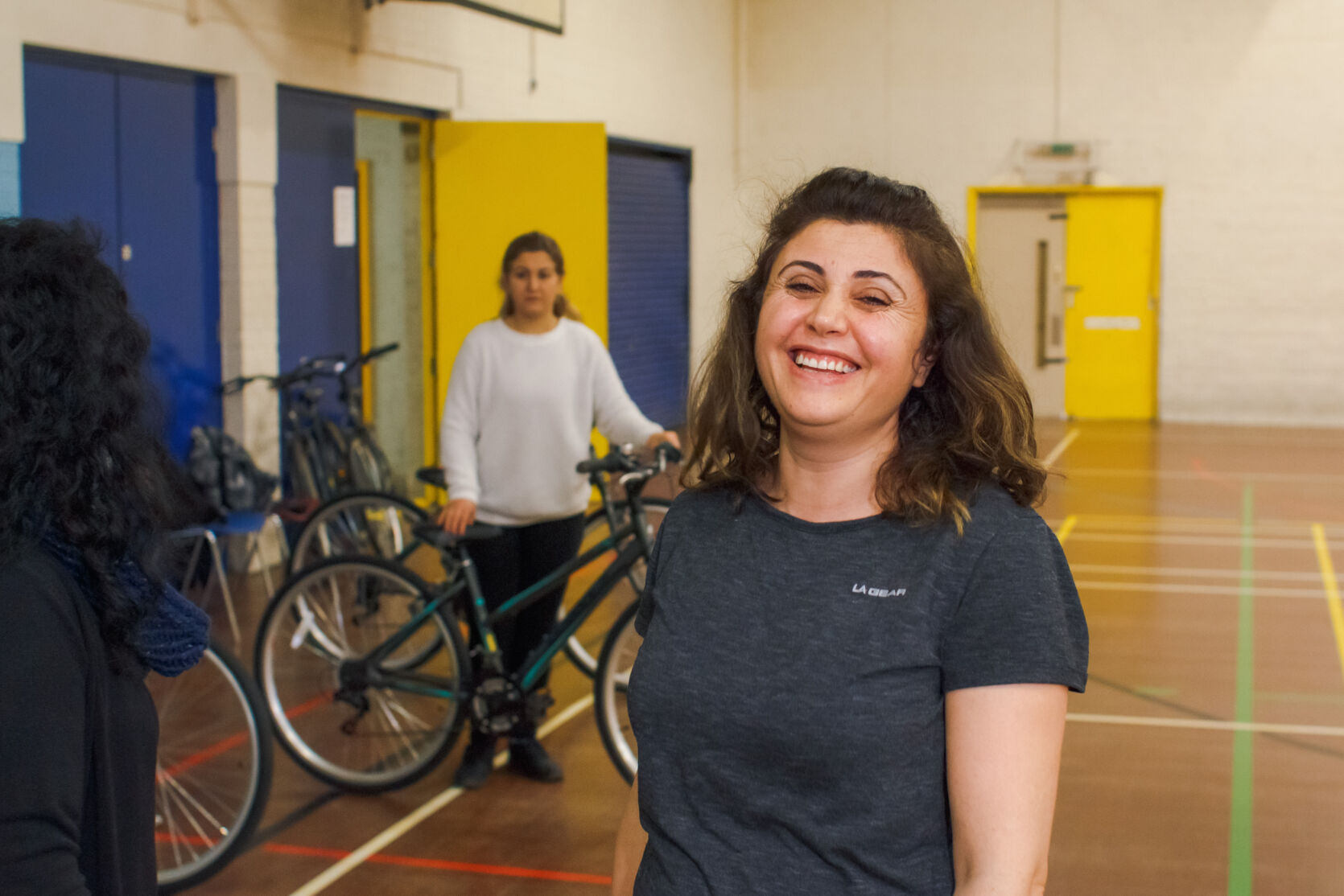 A women standing in a sports hall smiling at the camera. Behind her two other people holding bicycles - one is looking at the camera and the other has their back to us.