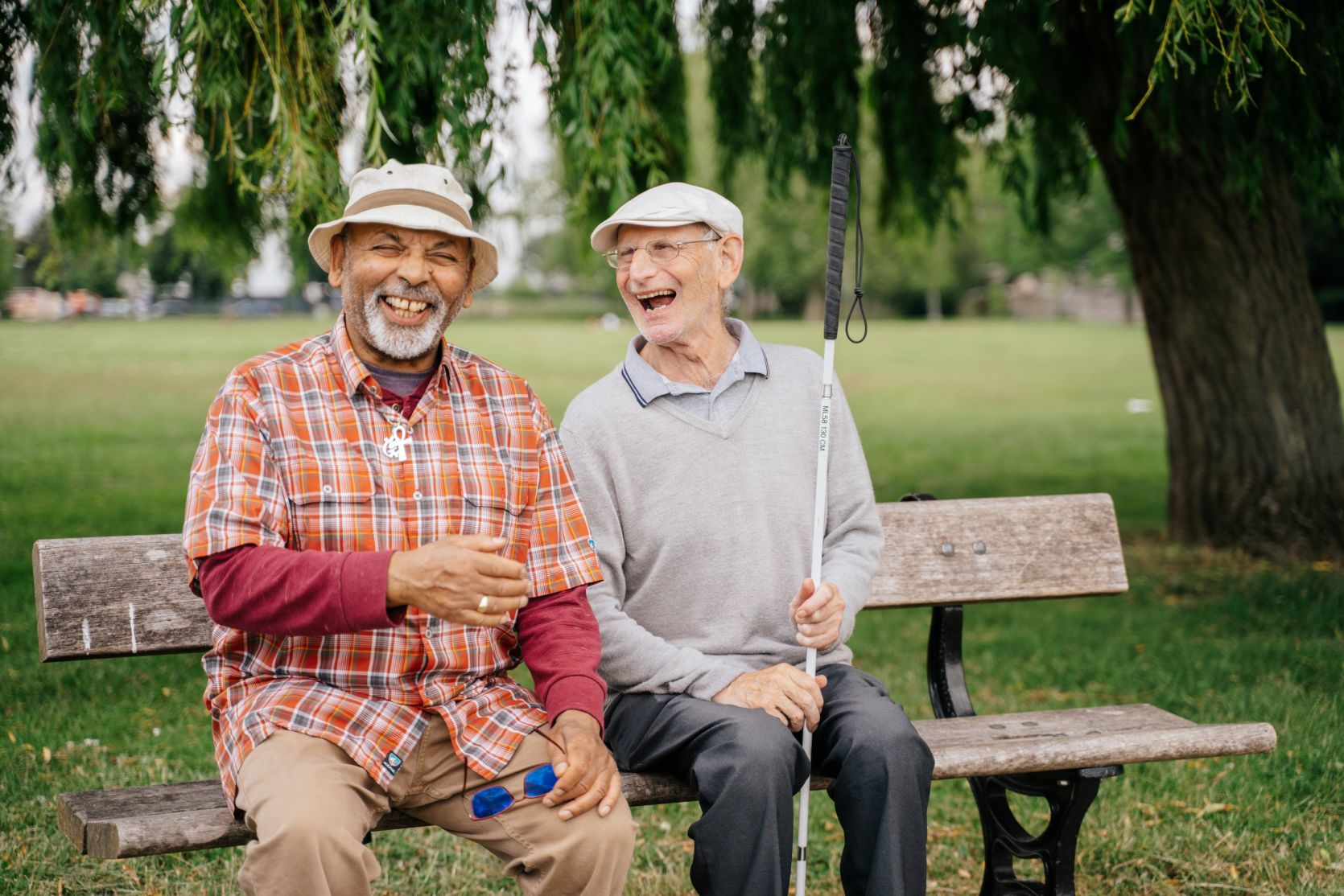 Two people laughing as they sit together on a park bench. They are both wearing hats while one has a pair of glasses and the other is carrying a white stick. Trees and a patch of grass can be seen behind them.