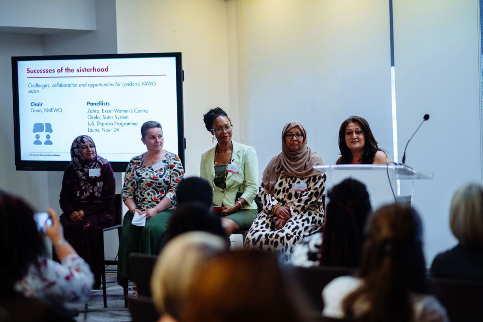 Panellists sit together in a line at the VAWG Grassroots Fund event. In the foreground you can see the back of the audience watching. In the background is a large screen featuring presentation slides.