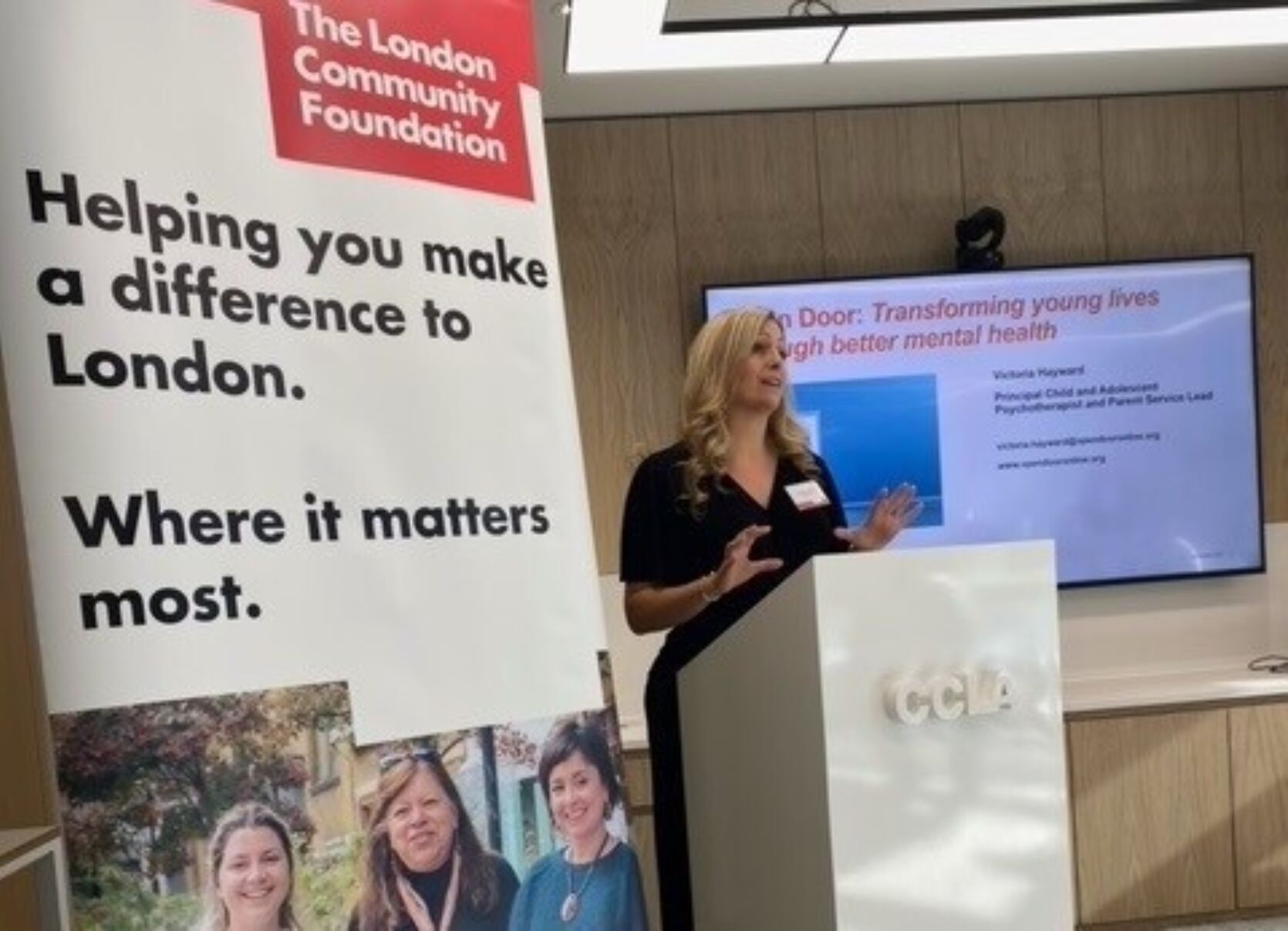 A person presenting at the Youth Futures Fund event. In the foreground is a London Community Foundation pull-up banner containing the words 'The London Community Foundation. Helping you make a difference to London. Where it matters most.' In the background is a large screen featuring some presentation slides.